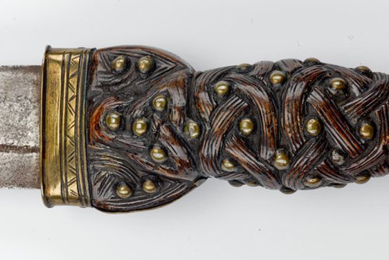 Close-up of an inicately carved dark brown wooden hilt of a dagger, with a bronze coloured metal band at the top edge.