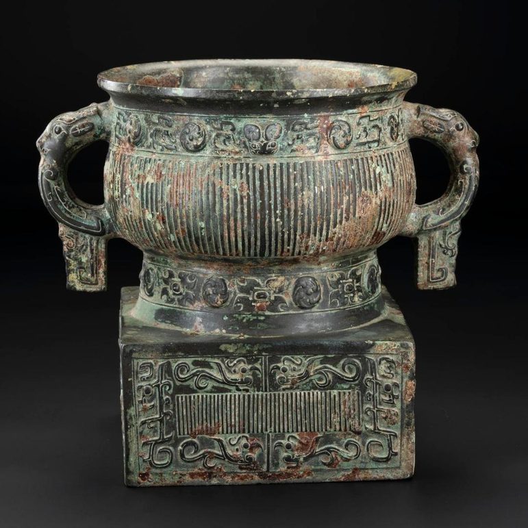 A large bronze cup with handles on both sides and a thick square base. It has engraved patterns of lines and dragons.