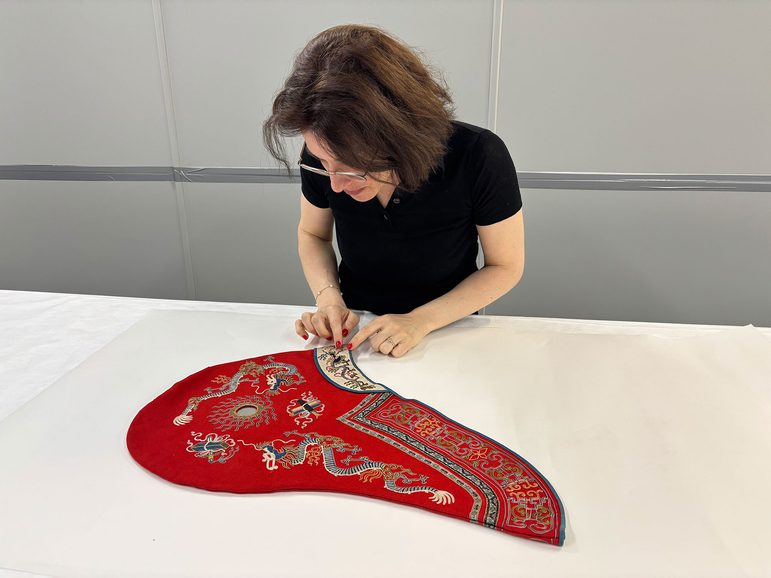 A historian examines an elaborate Chinese dress hood. The fabric is red and has embroidered designs of dragons.