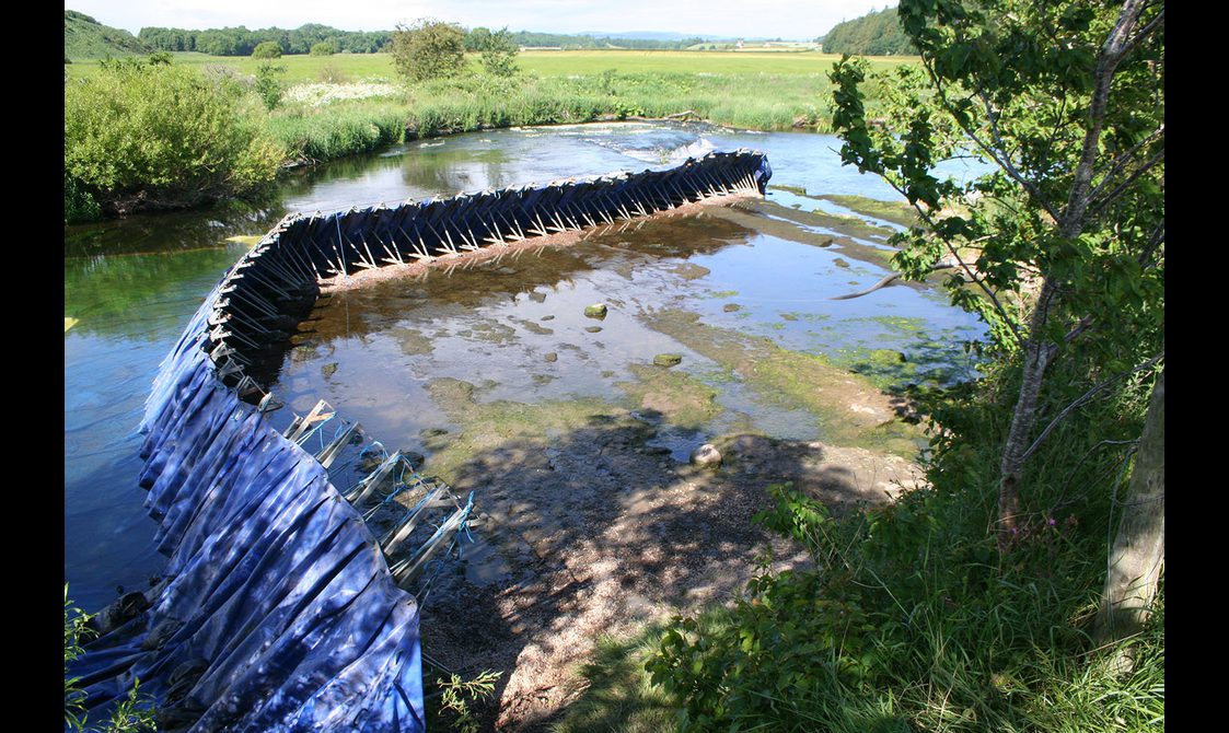 A curved dam made of steel polls and blue tarp running across a shallow river surrounded by green grass