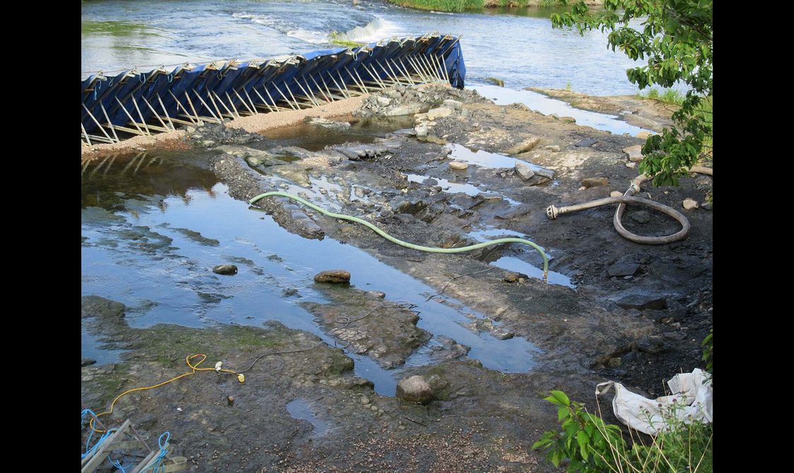 Green and grey hoses partially submerged in shallow water divided by a dam created with steel polls and blue tarp