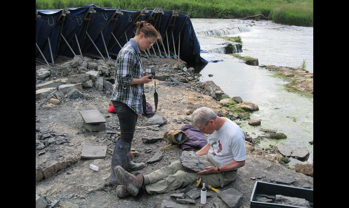 A woman in a checked shirt holding a camera standing beside a man in a white t-shirt sitting on a rocky ground examining a rock