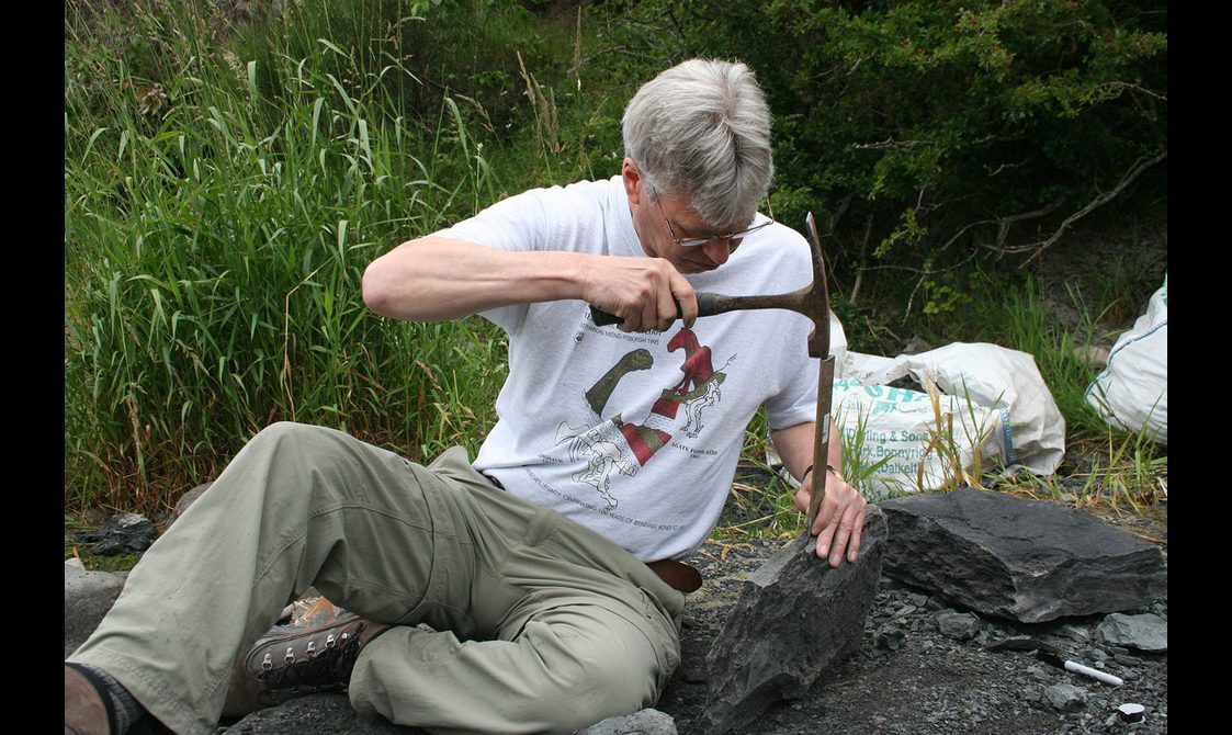A man with grey hair and glasses wearing a white tshirt and beige trousers sitting on a river side, splitting rocks with a chisel and hammer