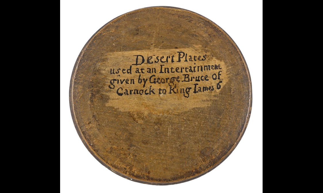 A round wooden object with black text that says Desert Plates used at an Intertainment given by George Bruce of Carnock to King James 6