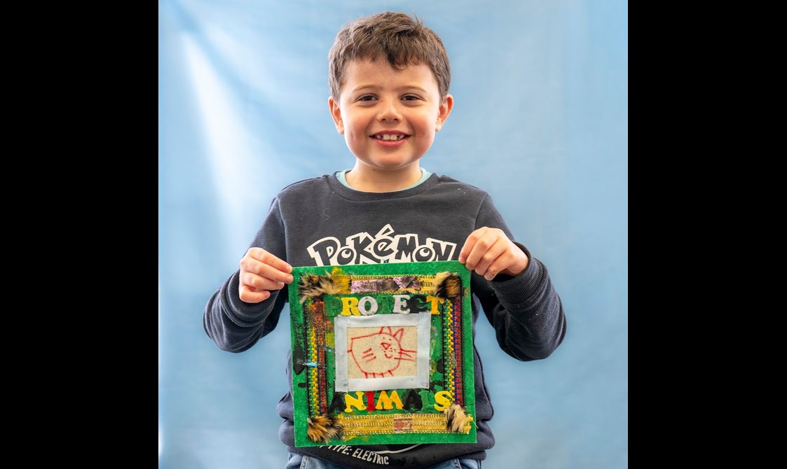A young person is smiling directly at the camera, holding up a fabric quilt scare that reads 'protect the animals'.