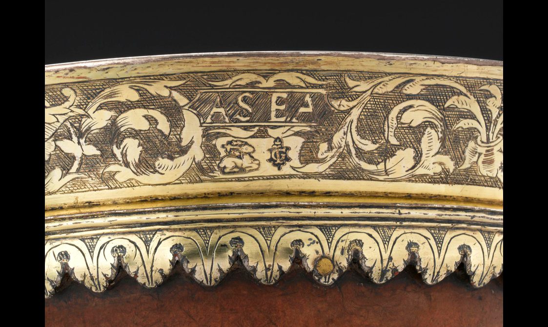 Closeup of the rim of a cup made from gold-coloured silver. The metal is engraved with the initials 'AS' and 'EA', and flanked by floral patterns.
