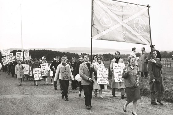 Black and white photo of a large group of young people walking and holding homemade signs with peaceful slogans.