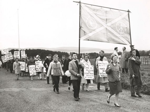 Black and white photo of a large group of young people walking and holding homemade signs with peaceful slogans.
