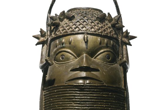A bronze sculpture of a face with large eyes with a lattice detailed headpiece. The sculpture has a thick base with coiled rings.