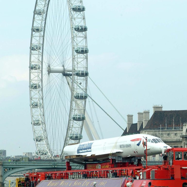 Part of a concorde airplane strapped on a large red truck in front of the London eye Ferris wheel