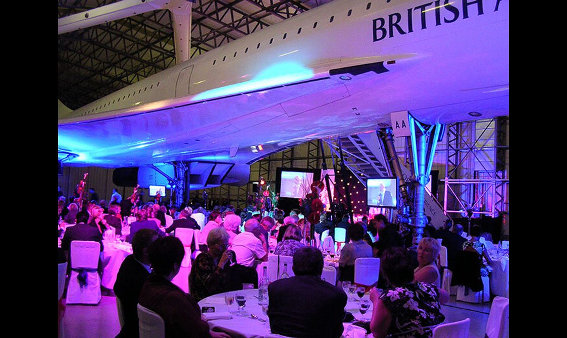 A group of people sitting down for dinner underneath the Concorde in a large aircraft hangar.