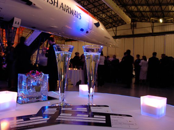 Two champagne glasses and candles on a table. A party full of people can be seen in the background under the nose of Concorde.