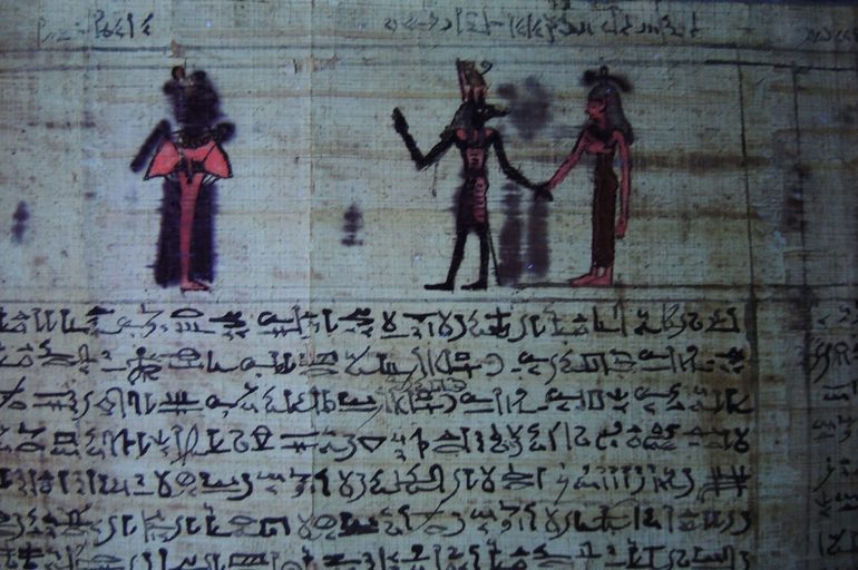 Detail of an ancient Egyptian papyrus with illustrations and hieroglyphs, shown under lighting that highlights a fluorescent pink colour on a figure.