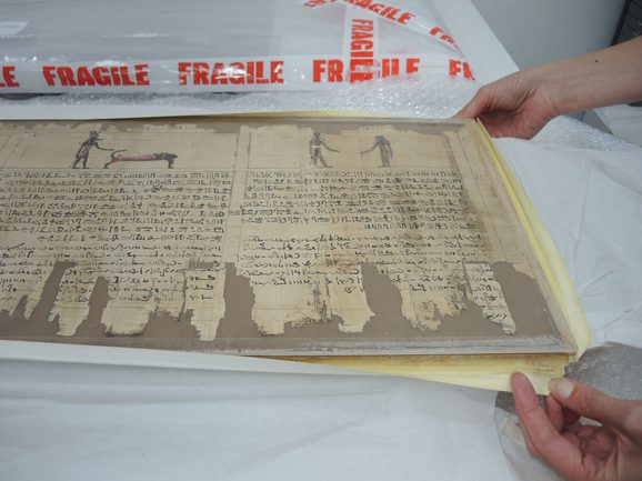 A yellowed, piece of paper with fragments missing featuring illustrations and hireographic text is mounted on brown cardboard. The edges of the cardboard are held by a pair of hands.