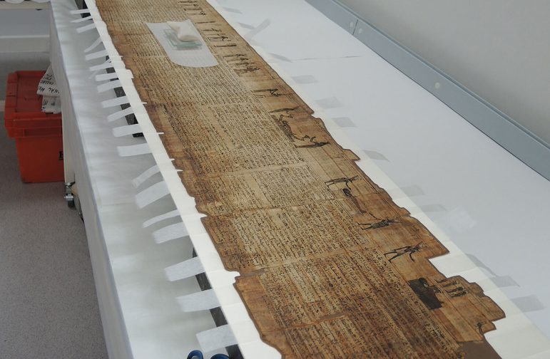 A yellowed ancient Egyptian scroll mounted and laid out on a long white table with tabs of tape sticking out from each side of the mount.