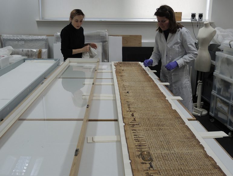 Two women examine a long anicent scroll laid out on a table in a lab setting.