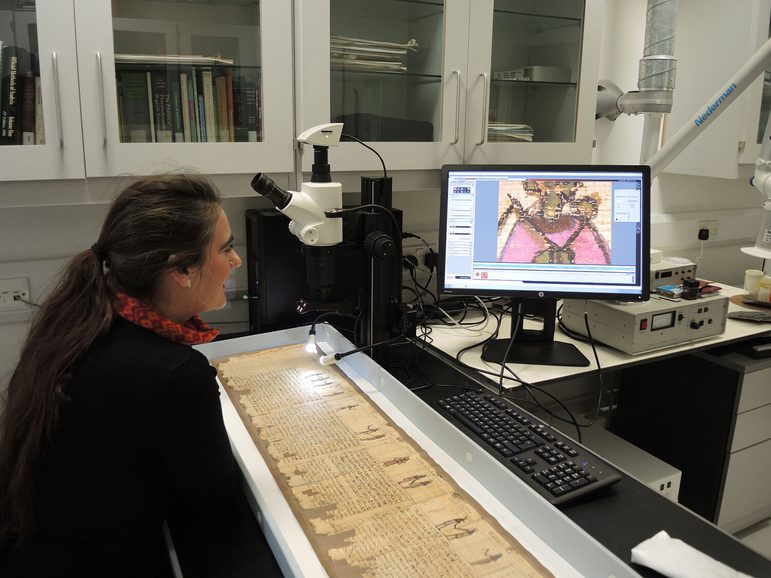 A waoman sits at a table in a lab setting with an ancient Egyptian scroll laid out in front of her. There is a microscope over the scroll with details being shown on a computer screen.