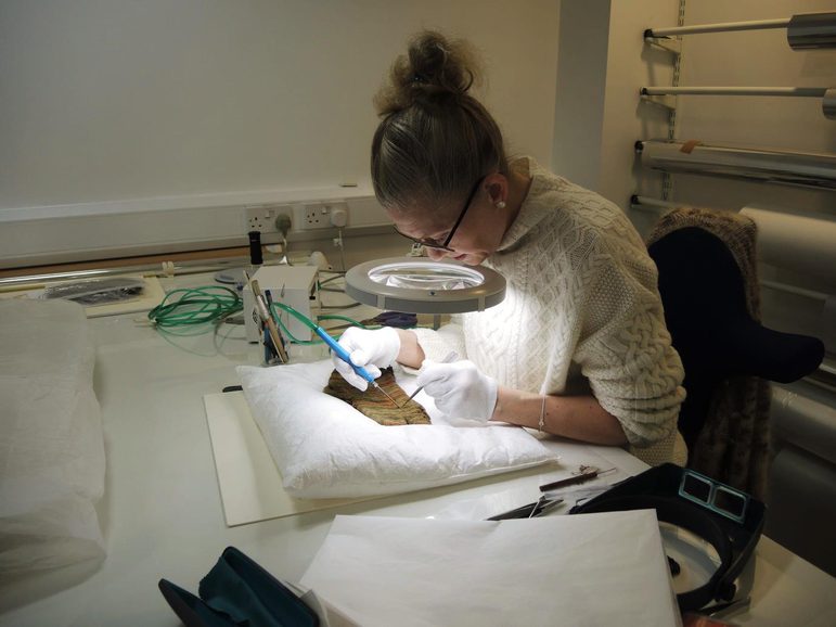 A woman in a cream knitted jumper delicately cleaning a textile sitting under a light and magnifying glass.