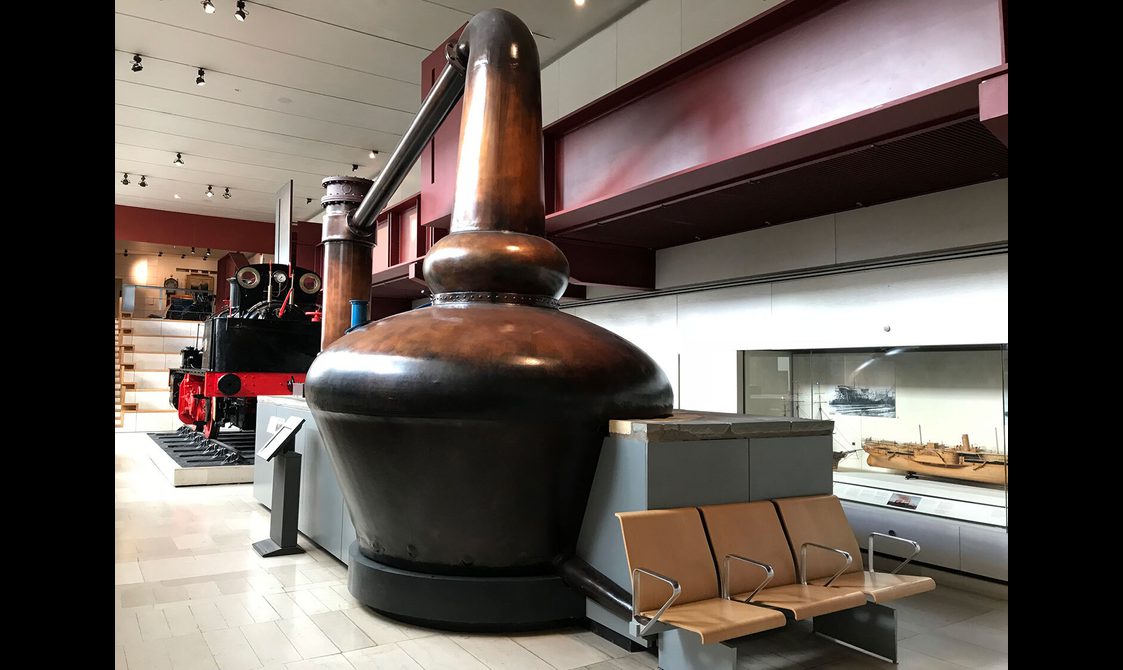 A museum setting with a large cylinder shaped canister that widens to the top with a hose attached to the top.