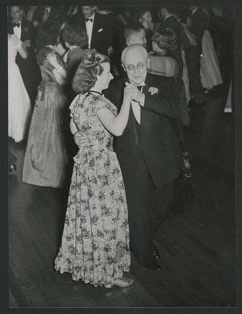 A black and white photograph of a man wearing a suit, dancing with a woman surrounded by others on the dancefloor.