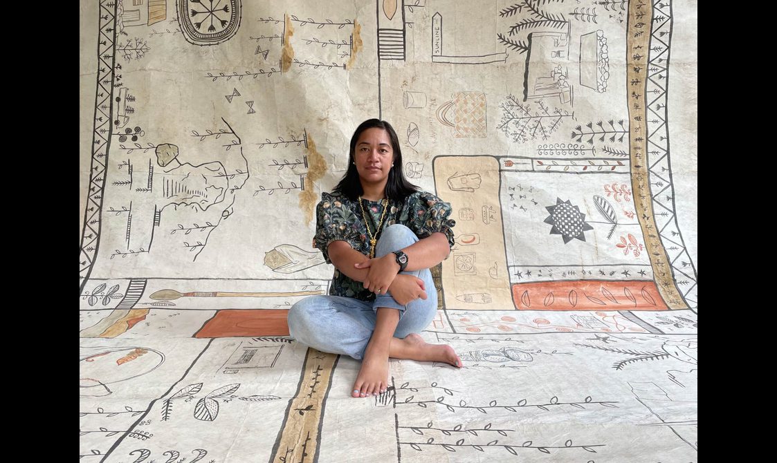 A woman sits holding her legs crossed on a large piece of cloth which extends up behind her. It is covered in hand printed or painted illustrations of leaved and plants.