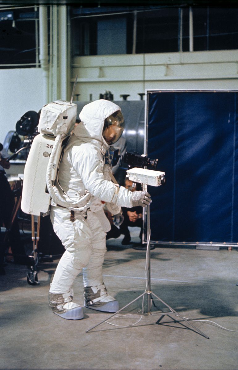 An astronaut in an astronaut suit stands in a studio holding a camera on a tripod.