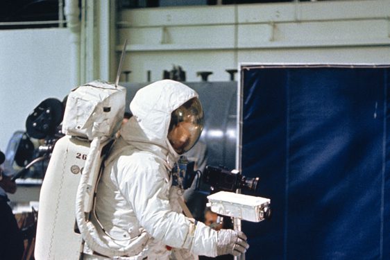 An astronaut in an astronaut suit stands in a studio holding a camera on a tripod.