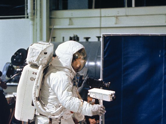 An astronaut in an astronaut suit stands in a studio holding a camera on a tripod.