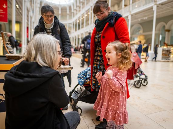 A family in a large museum gallery. A member of staff is kneeling down to talk to a child who is very excited.