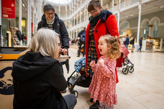 A family in a large museum gallery. A member of staff is kneeling down to talk to a child who is very excited.