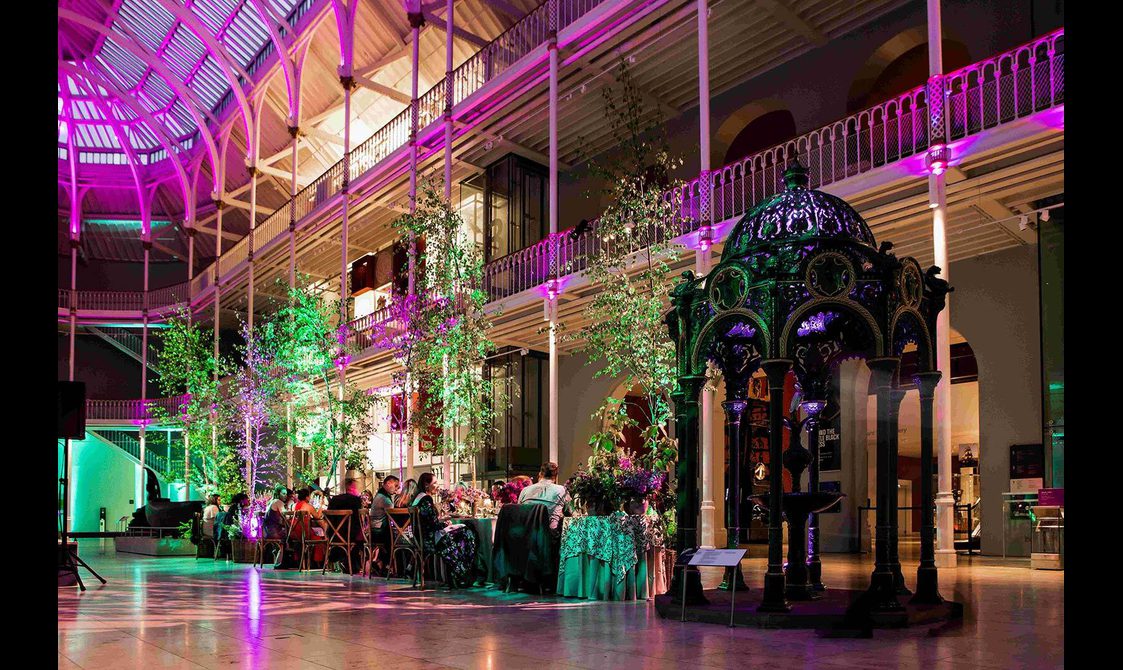 A dinner reception in a large multi-level museum gallery. There are trees lining the dinner table and purple lights illumintaing the space.