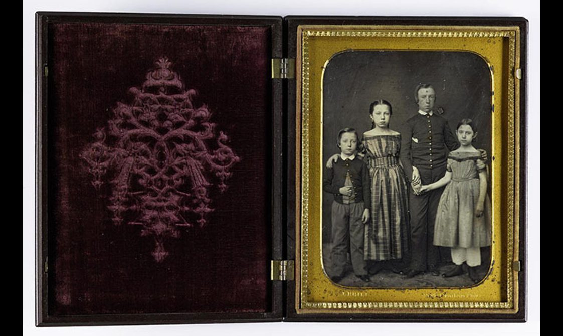 A photo album open with a purple velvet cover to the left with a black and white image of a man and a woman with two children, a boy and a girl, in Victorian dress. The image has a gold frame.