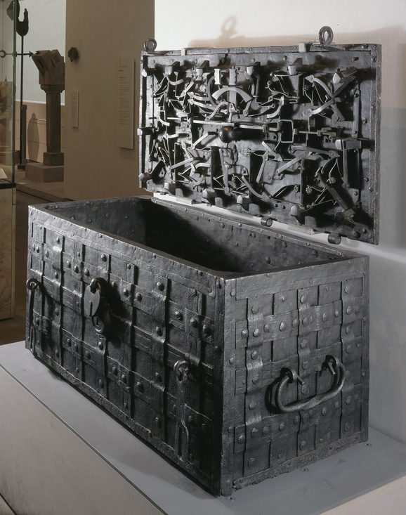 A large open iron chest with a grid pattern on its surface and an intricate lock mechanism and design on the underside of the lid. The chest sits on a grey plinth in a museum setting.