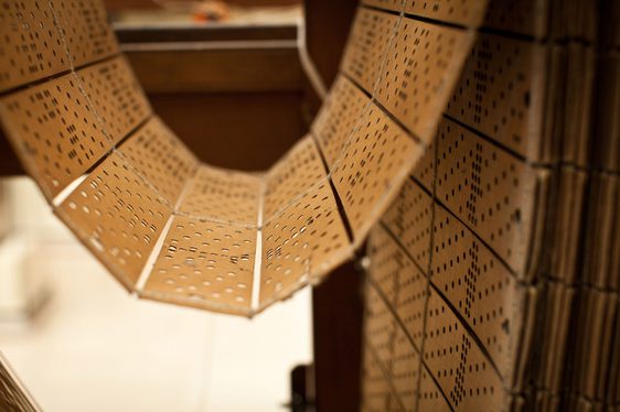A series of light brown cards with patterns of holes punched into them sticthed together and hanging looped on a wooden frame