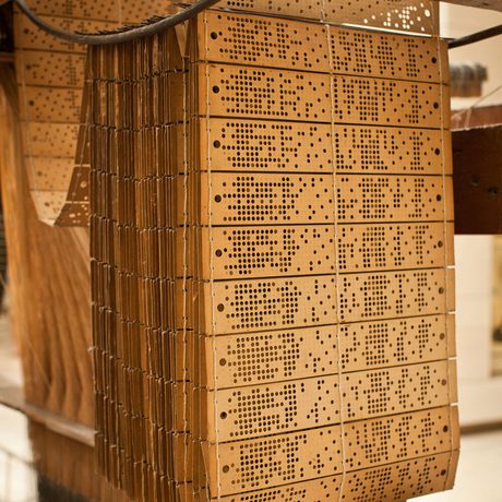 various sets of dark yellow cards with patterns of hole punched into them are sticthed together, hanging on a wooden loom