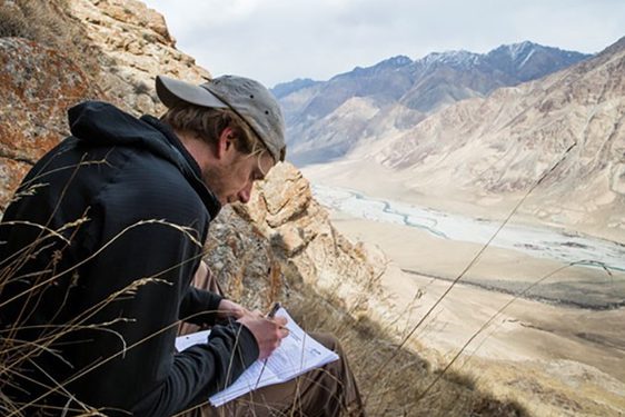 A man sits on the side of a hill overlooking a dry brown valley. He is writing on a piece of paper.