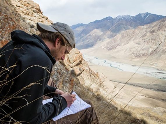 A man sits on the side of a hill overlooking a dry brown valley. He is writing on a piece of paper.