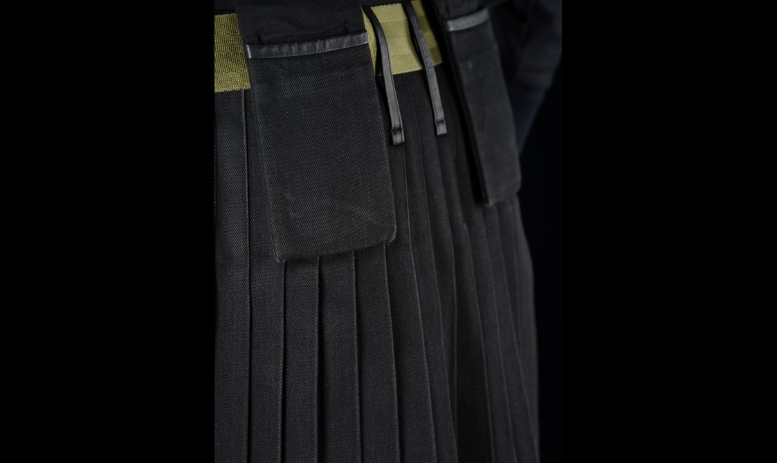 Detail of a black denim kilt showing pockets, pleats, and a green belt.