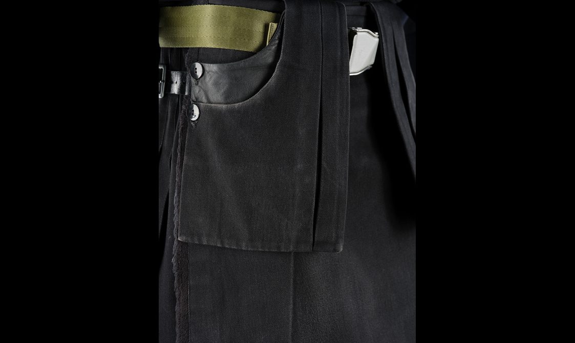 Detail of front pockets with a woven green belt on a black denim kilt.