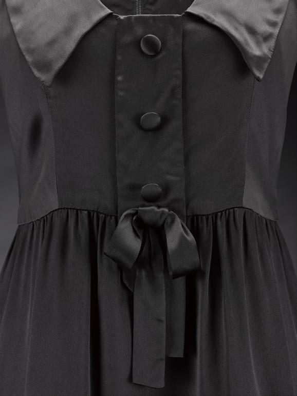Detail of a black satin dress with large collar, three buttons down the front, and a small bow.