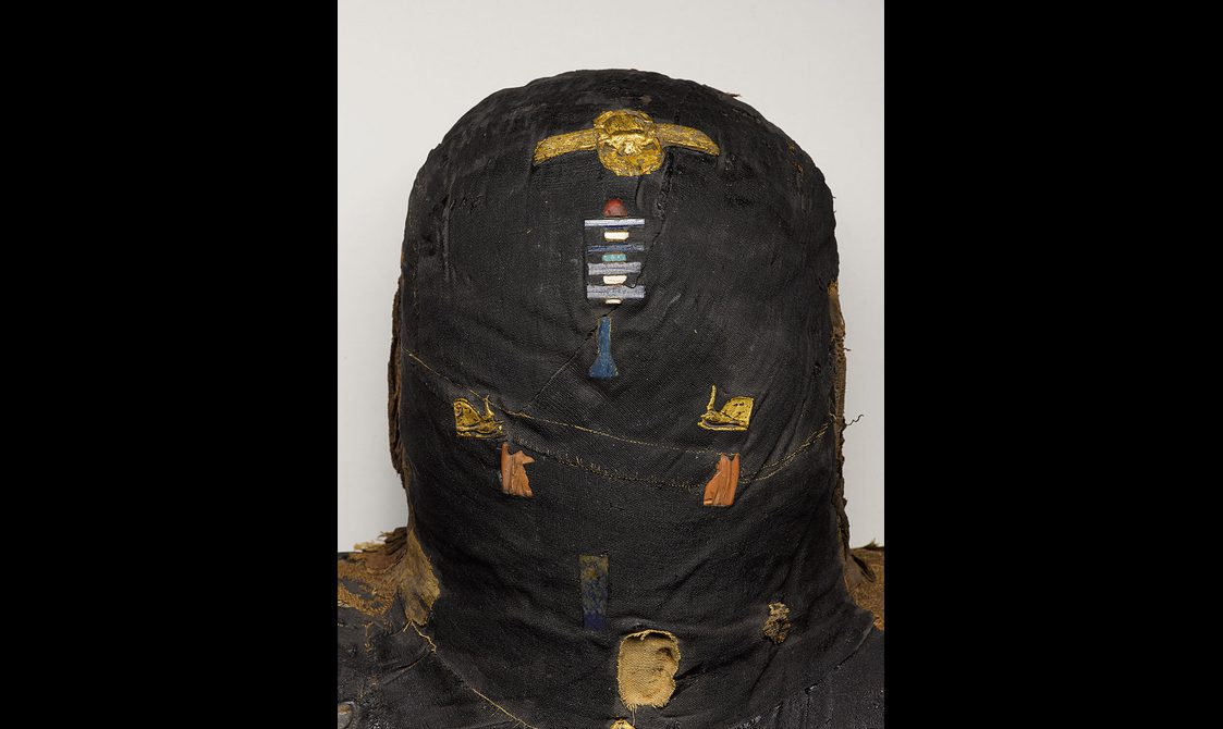 Detail of the head of a mummified woman wrapped in blackened bandages with amulets and decorative objects embedded on the surface.