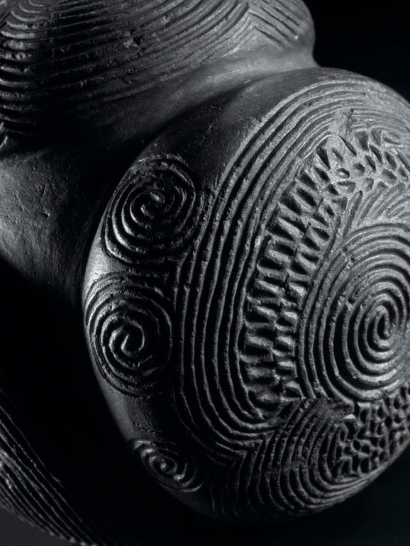 A close up detail of a disk ball with horizontal and spiral carvings set against black background.