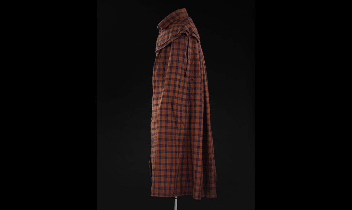 A side view of a mannequin wearing a long brown, navy, and red tartan cloak