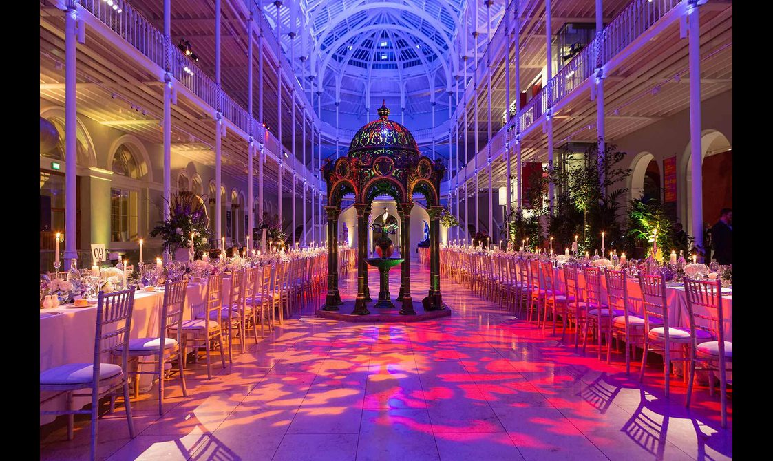 Long tables lined with chairs in a multi-level gallery space with balconies running around the outside. It is lit in pinks, blues, and purples.