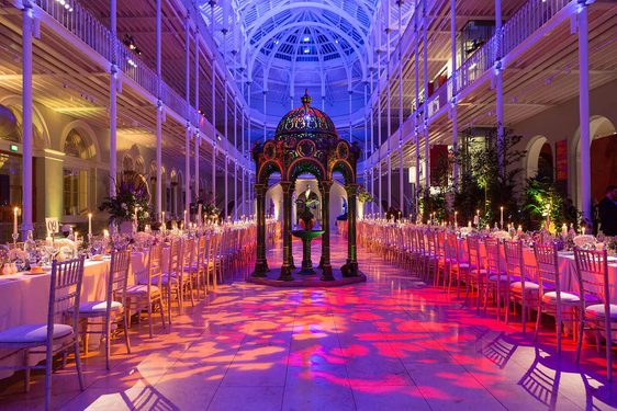 Long tables lined with chairs in a multi-level gallery space with balconies running around the outside. It is lit in pinks, blues, and purples.