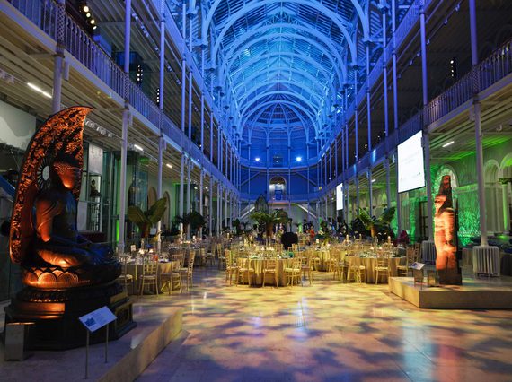 A large multi-level atrium set up for an evening dinner event. Round tables are set up for dinner and the space is illuminated with different coloured lights.