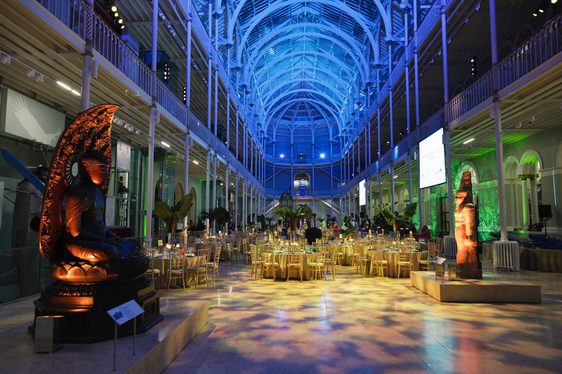 A large multi-level atrium set up for an evening dinner event. Round tables are set up for dinner and the space is illuminated with different coloured lights.
