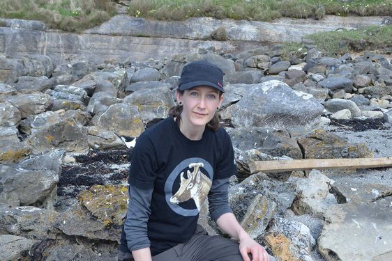 A woman in dark t-shirt, trousers, and baseball cap kneeling on a rocky landscape beside a fossilised dinosaur bone