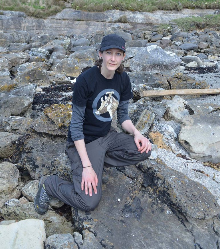A woman in dark t-shirt, trousers, and baseball cap kneeling on a rocky landscape beside a fossilised dinosaur bone