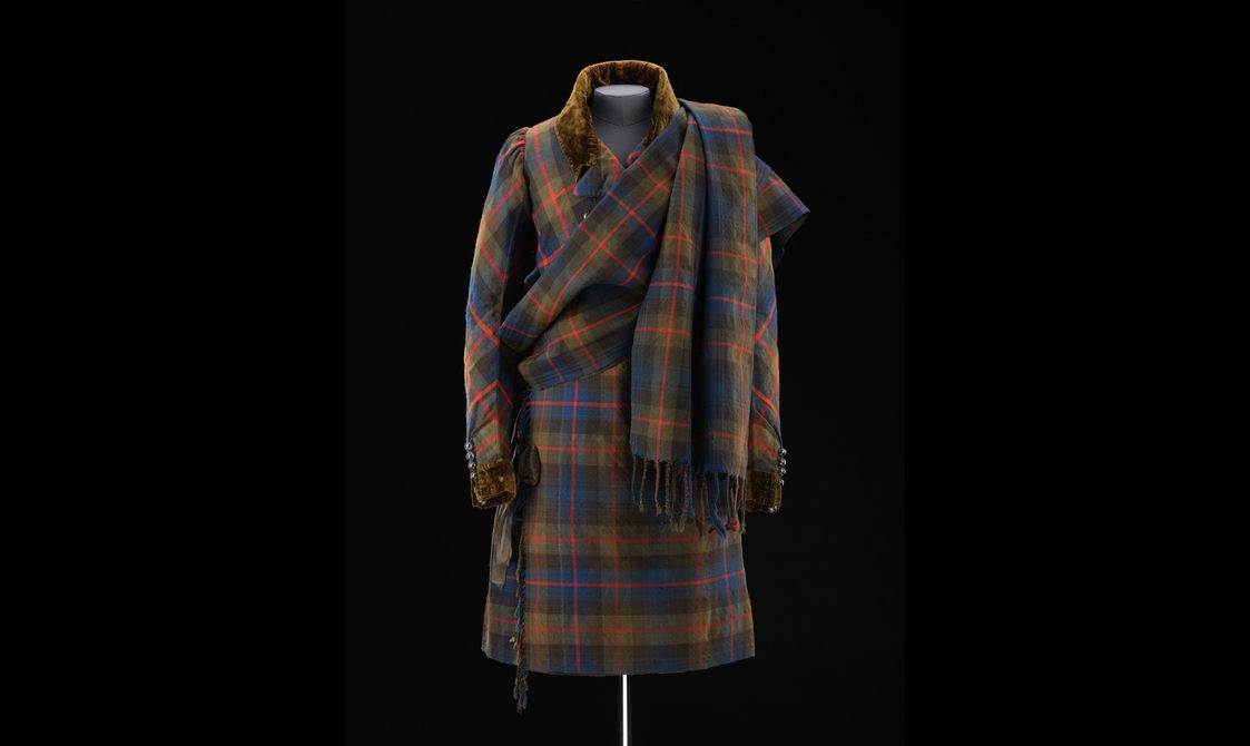 A mannequin wearing a navy, green, and red tartan kilt with a matching jacket and draped shawl.